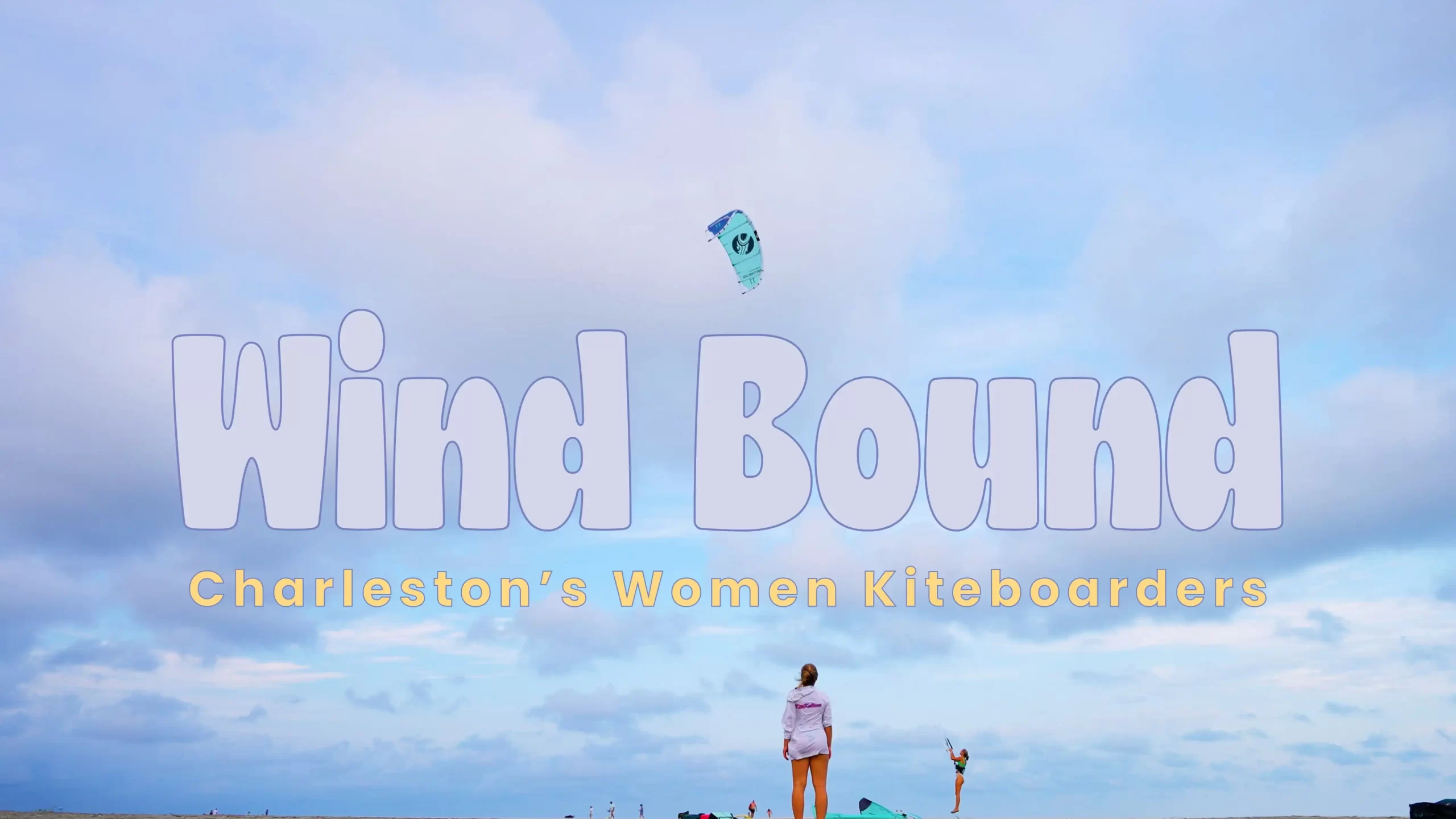 A woman stands on a beach looking up at a large kite in the blue sky.