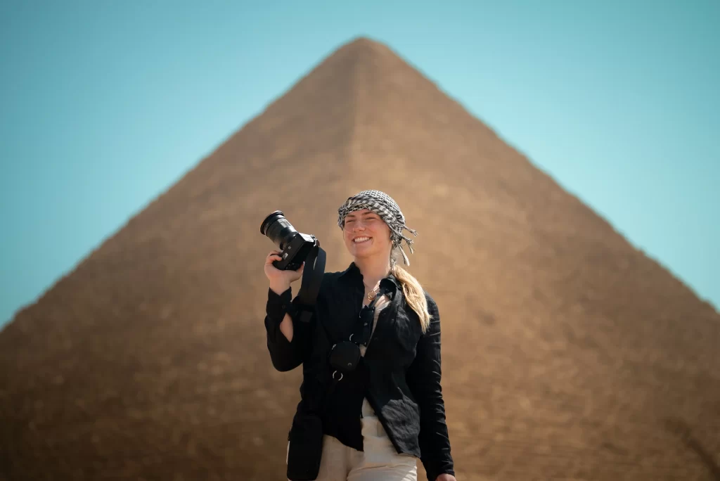 Person in black shirt and patterned headscarf holding a camera in front of a large pyramid.